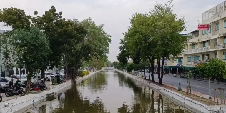 A straight, walled canal with calm, greenish water runs towards a small bridge in the distance under an overcast sky. Mature trees line both banks. Motorbikes and cars are parked along the left side, and a road with a row of low shophouses runs along the right.