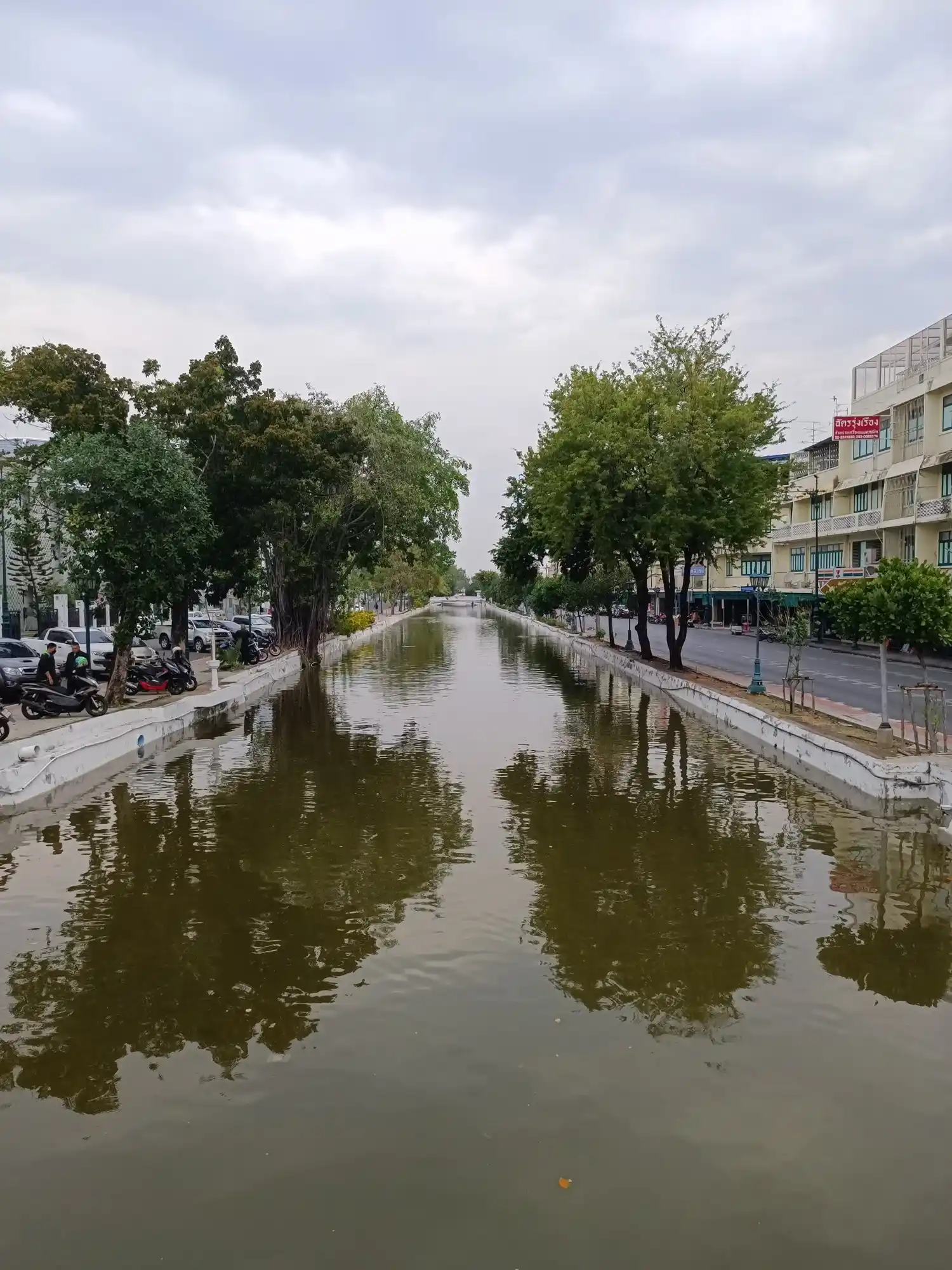A straight, walled canal with calm, greenish water runs towards a small bridge in the distance under an overcast sky. Mature trees line both banks. Motorbikes and cars are parked along the left side, and a road with a row of low shophouses runs along the right.