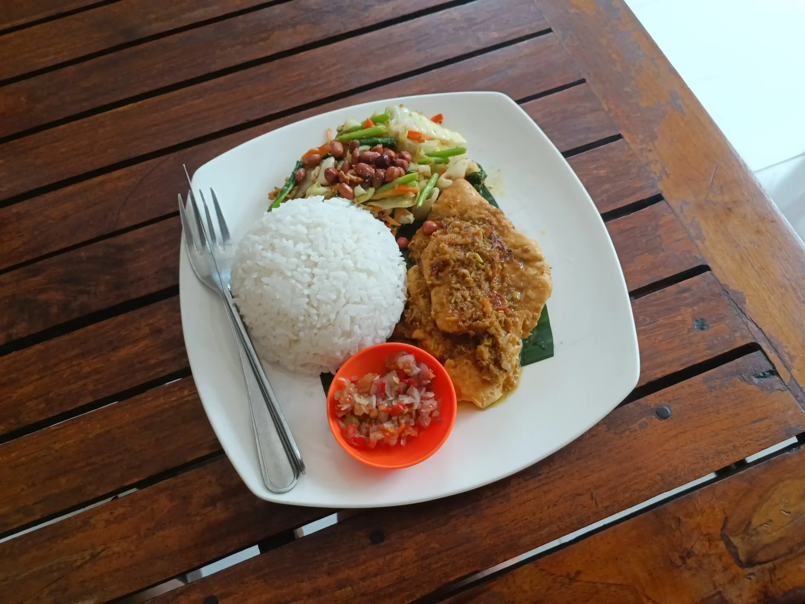 Ayam Betutu khas Bali disajikan dengan nasi putih, sayuran tumis, kacang tanah dan sambal di atas piring putih.