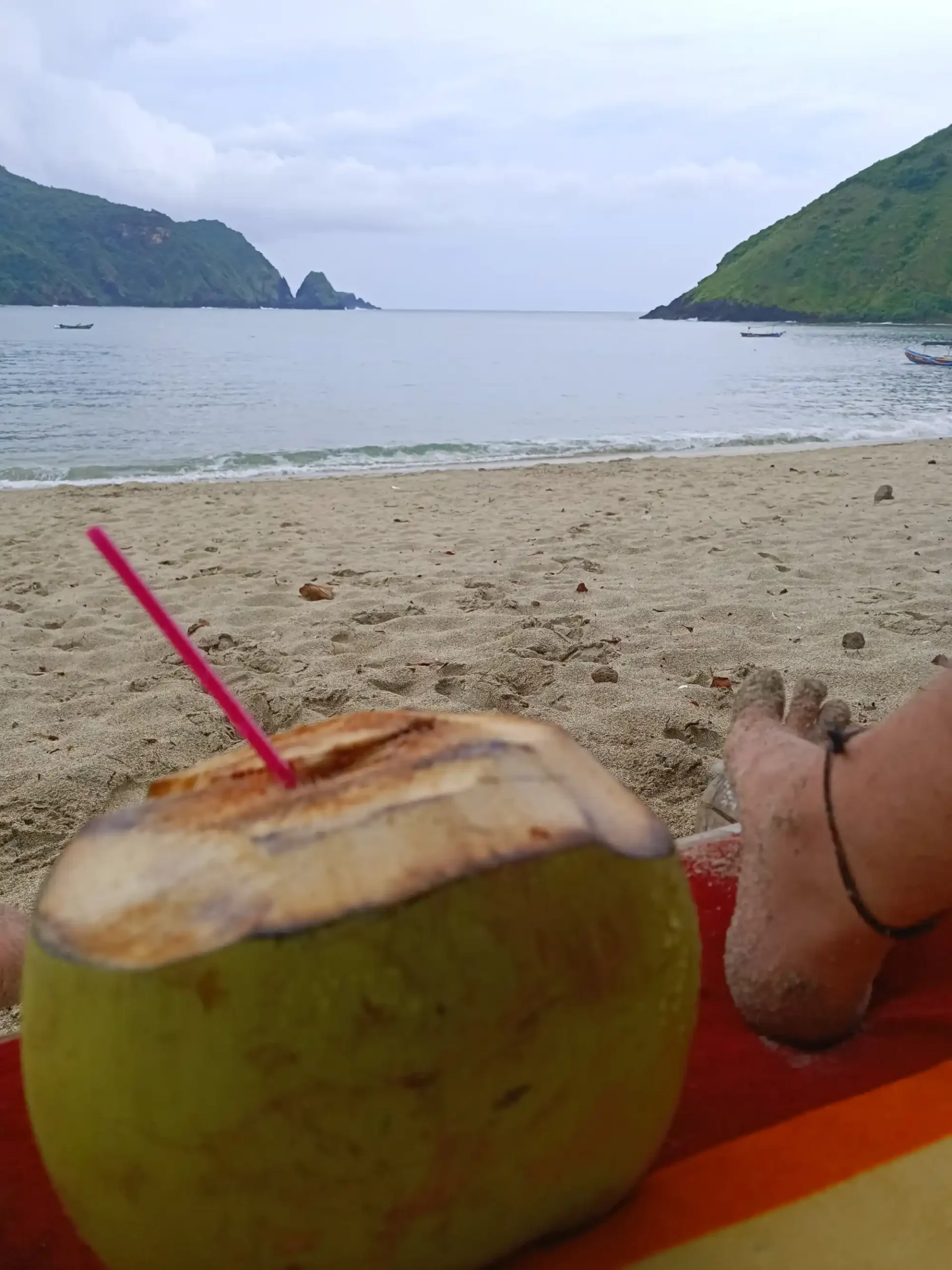 Coconut drink with a pink straw on the sand at Mawun Beach, with green hills and calm sea in the background.