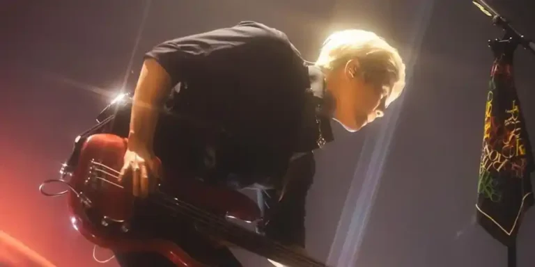 A member of The Rose playing bass guitar on stage while leaning forward beneath warm concert lights.