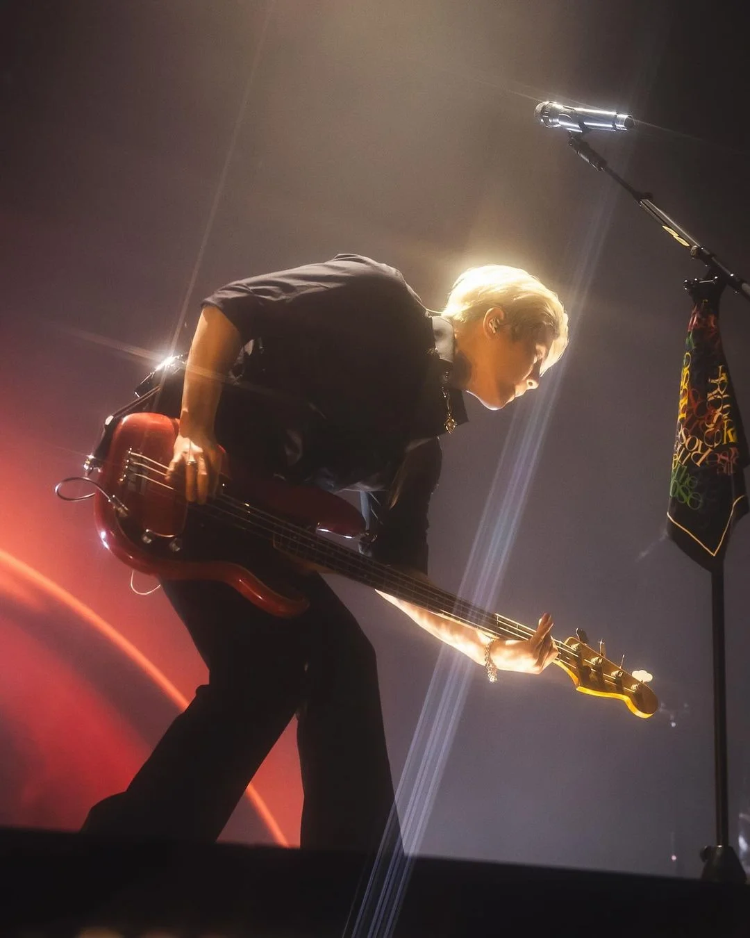 Anggota The Rose memainkan gitar bass di atas panggung sambil membungkuk di bawah cahaya konser.