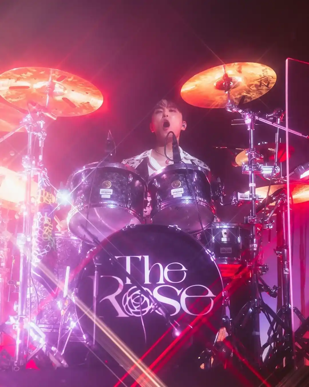 Anggota The Rose memainkan drum di atas panggung dengan cahaya konser merah dan merah muda.