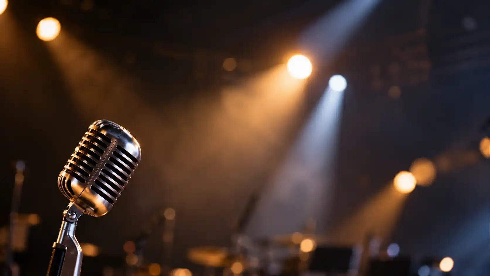 Mikrofon vintage di atas panggung gelap dengan sorotan lampu amber dan putih yang membentuk latar konser yang buram.