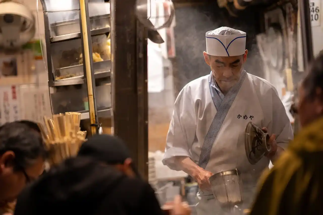 Japanese chef preparing food at a busy restaurant in Tokyo, wearing a traditional white chef’s uniform and hat.