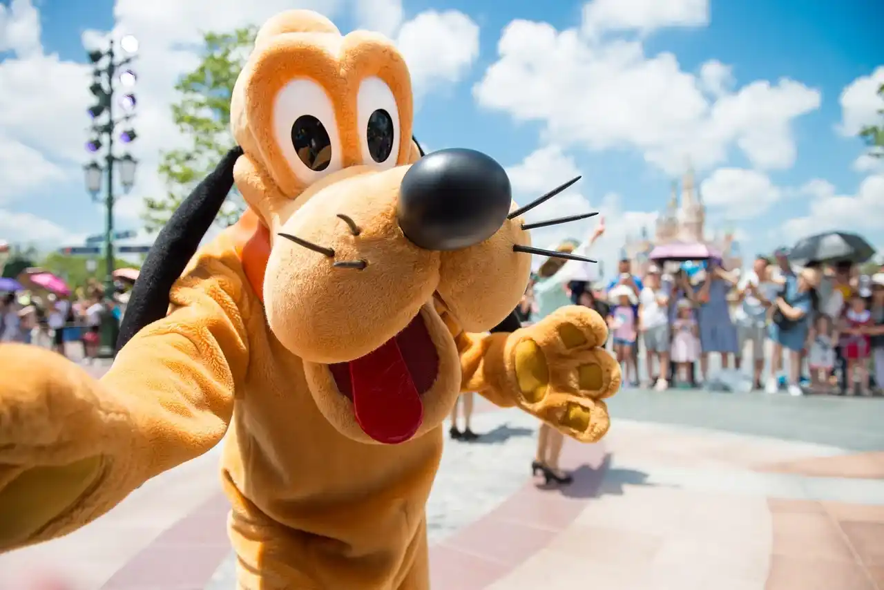 Pluto character mascot greeting visitors at Tokyo Disneyland, with guests gathered in the sunny outdoor park behind him.