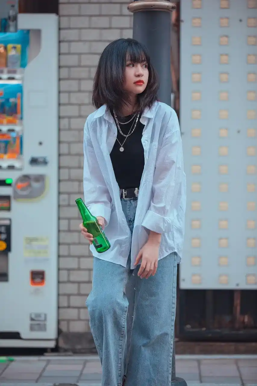 Young woman in Tokyo wearing a casual layered outfit and holding a green bottle beside a vending machine on a city street.