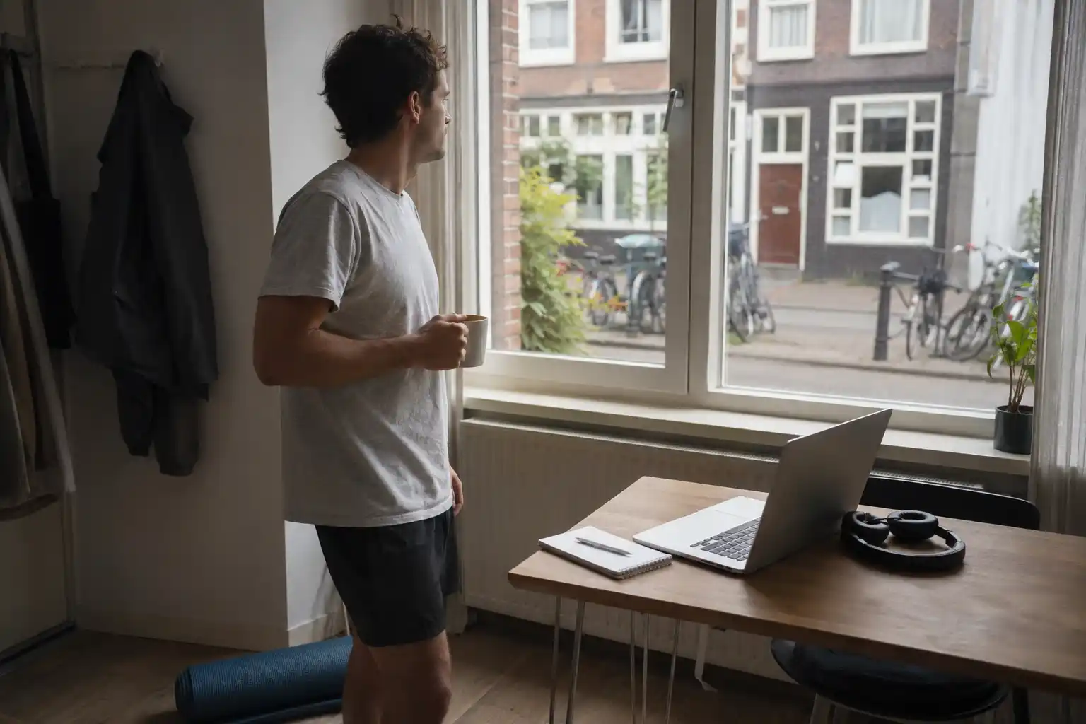 Male digital nomad drinking coffee beside his laptop after a morning workout in an Amsterdam apartment.
