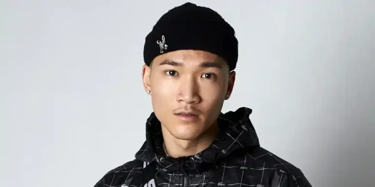 Young man wearing a black patterned hooded jacket, black trousers, black beanie and a black crossbody bag.