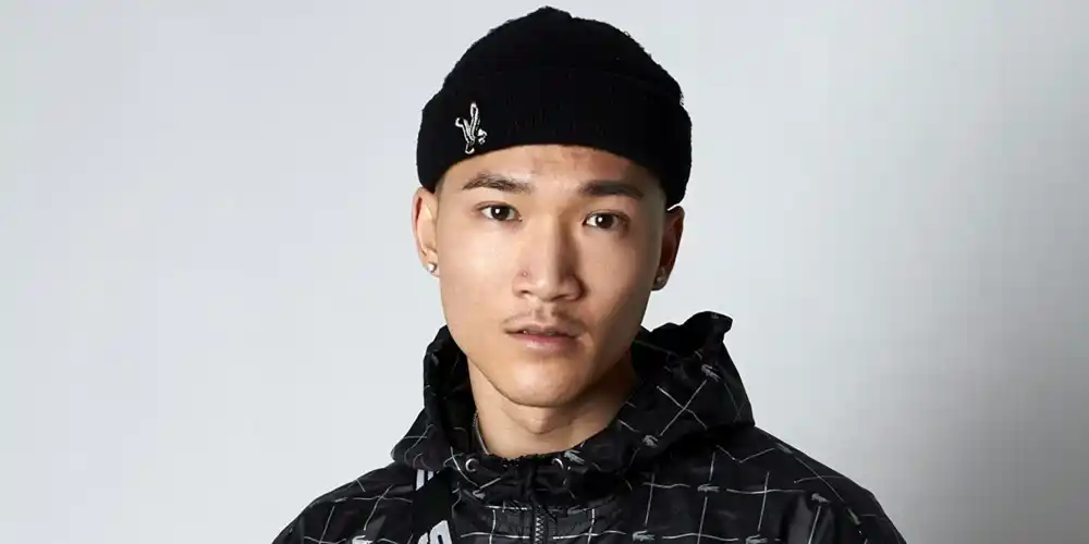 Young man wearing a black patterned hooded jacket, black trousers, black beanie and a black crossbody bag.