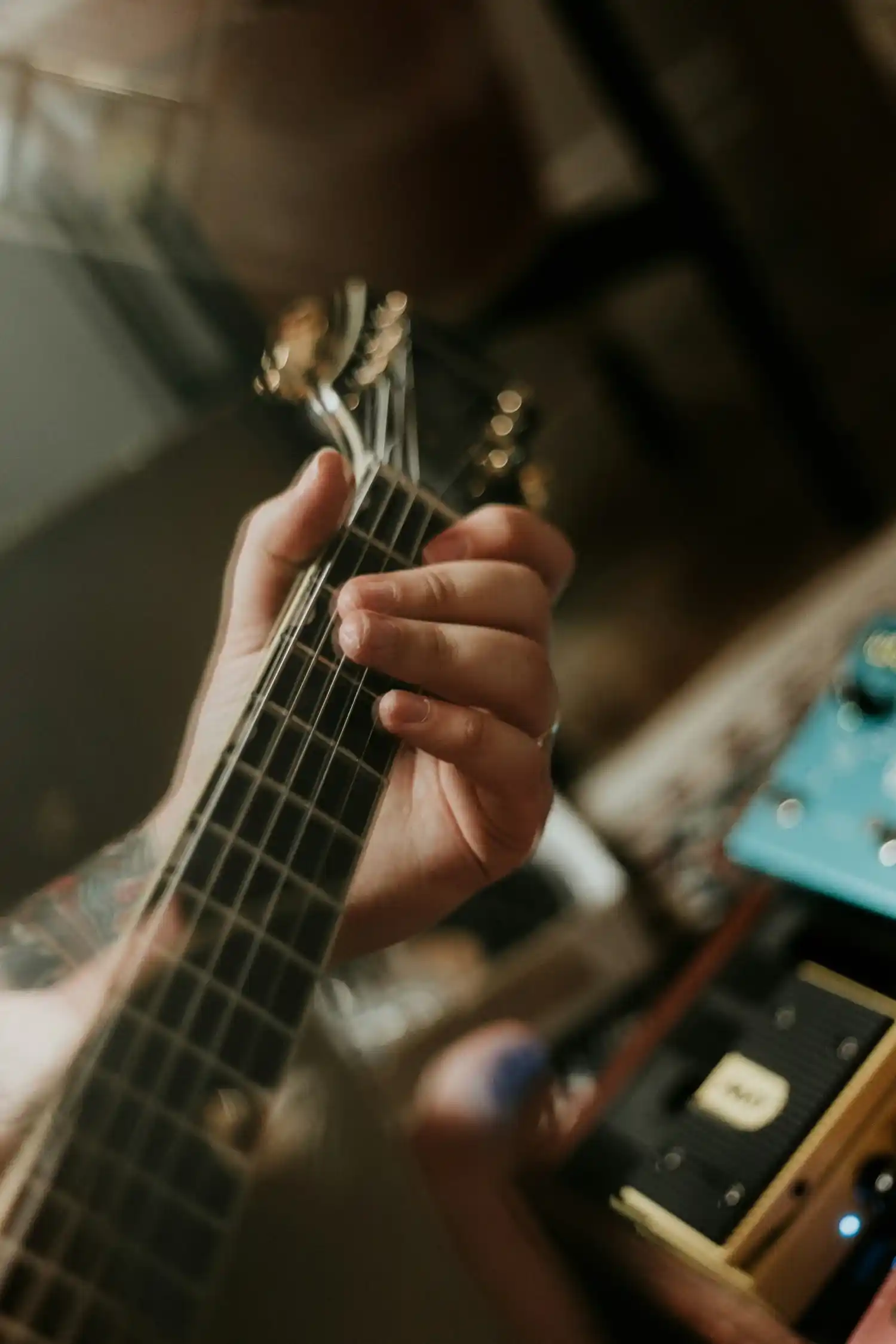 Close-up of a musician’s hand playing chords on an electric guitar, with guitar pedals visible in the background.