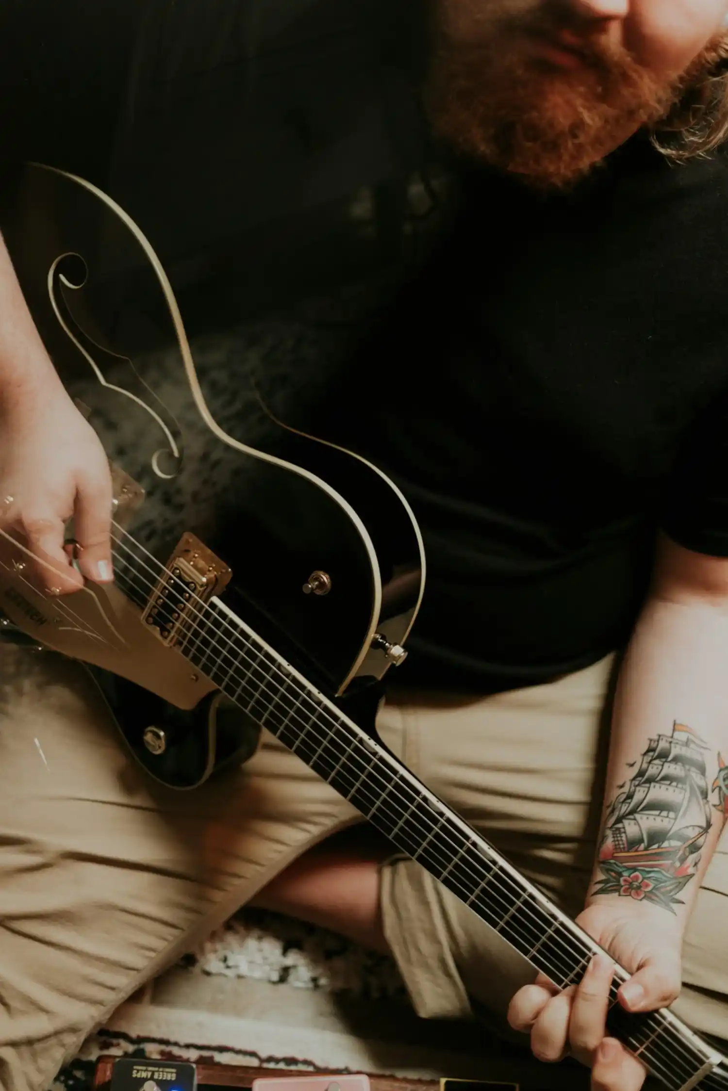 Bearded musician with a tattooed arm playing a black electric guitar while seated indoors.