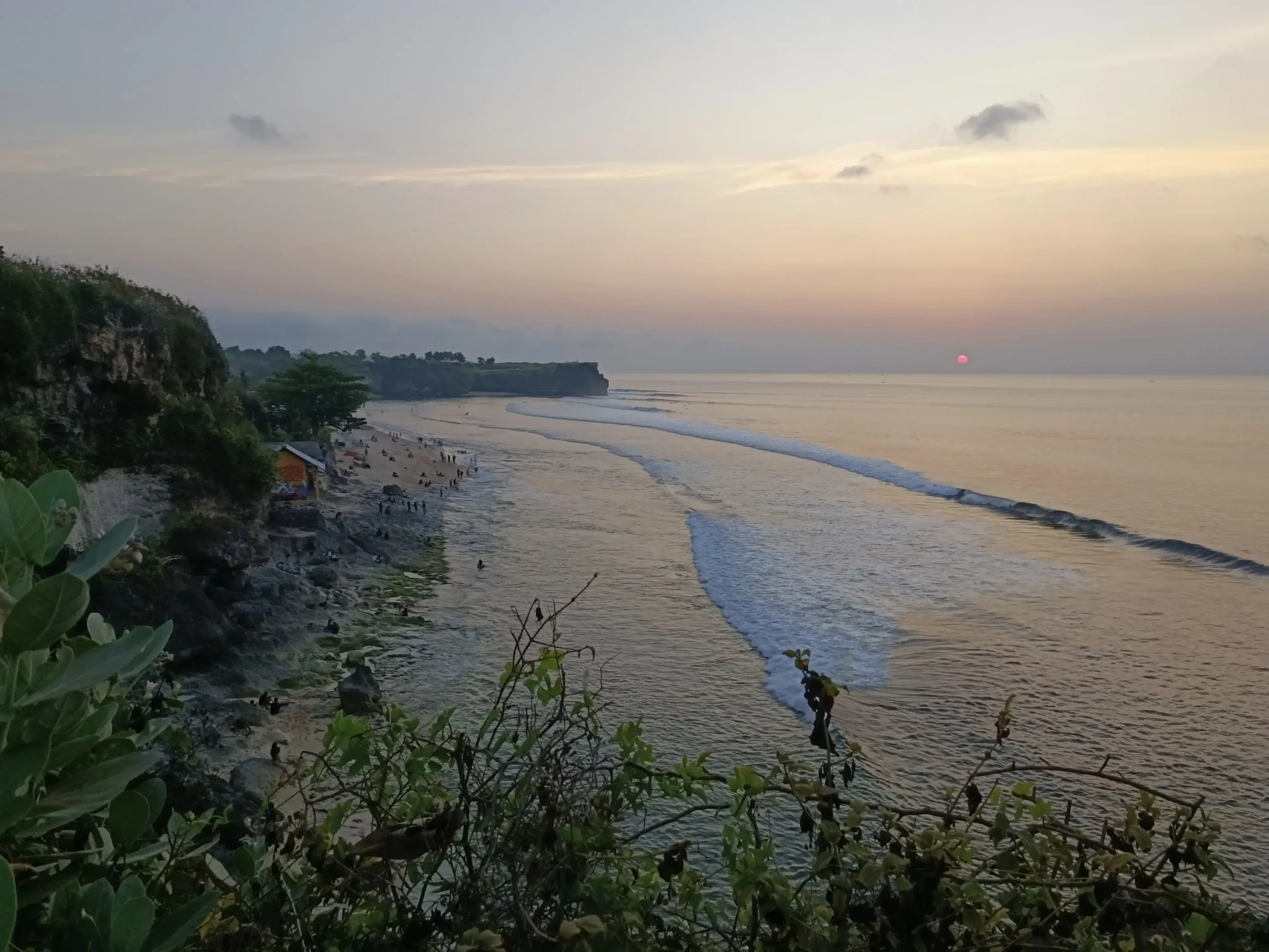 Matahari terbenam di Pantai Balangan, Bali, dengan ombak lembut menuju pantai, tebing berbatu yang ditumbuhi tanaman hijau dan orang-orang di sepanjang pantai.