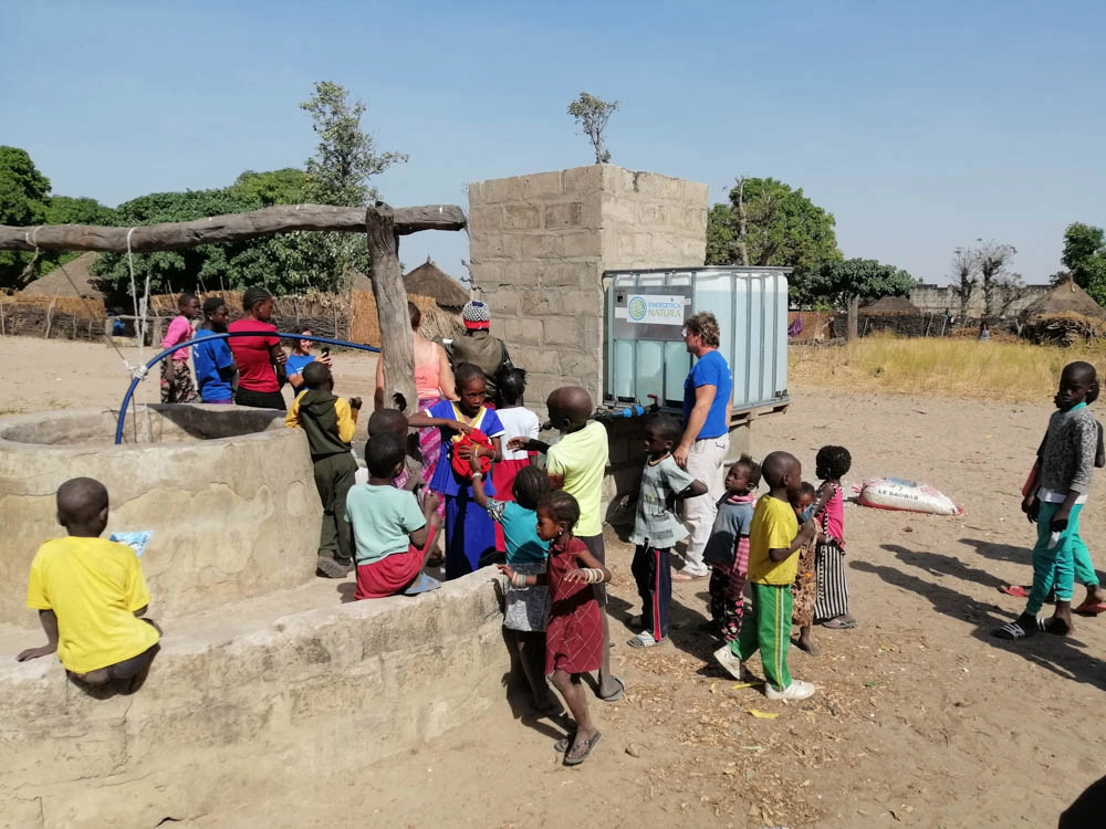 Children and adults gather around a community water well at Sandicoly Ecofarm in Senegal, beside a water storage container and traditional village huts.