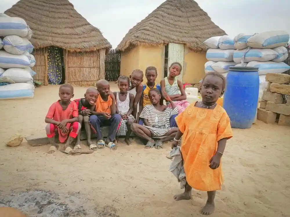 Children gather in a village at Sandicoly Ecofarm in Senegal, surrounded by traditional thatched-roof huts, sacks of goods and a blue water barrel.