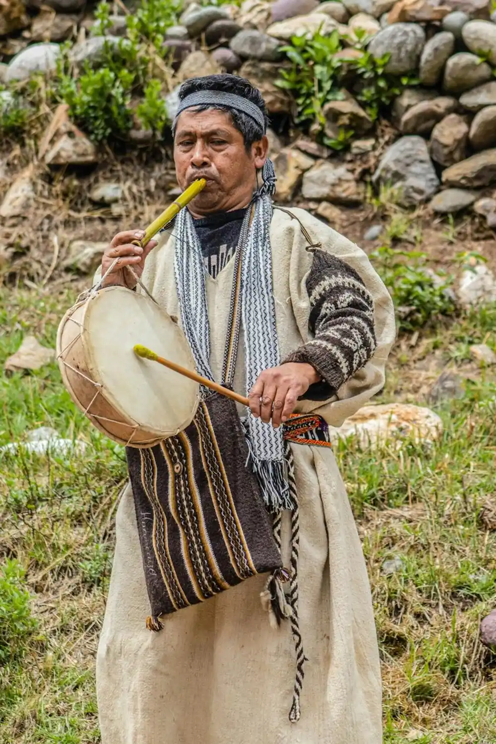 Man in traditional Andean clothing playing a flute and drum in a rural mountain setting in Peru.