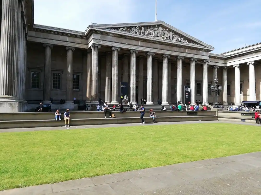 Exterior of the British Museum in London with classical columns and visitors sitting outside.