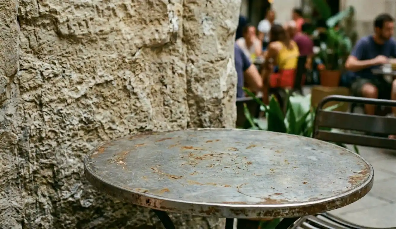 A worn metal café table beside a textured wall in Barcelona with blurred people sitting in the background.