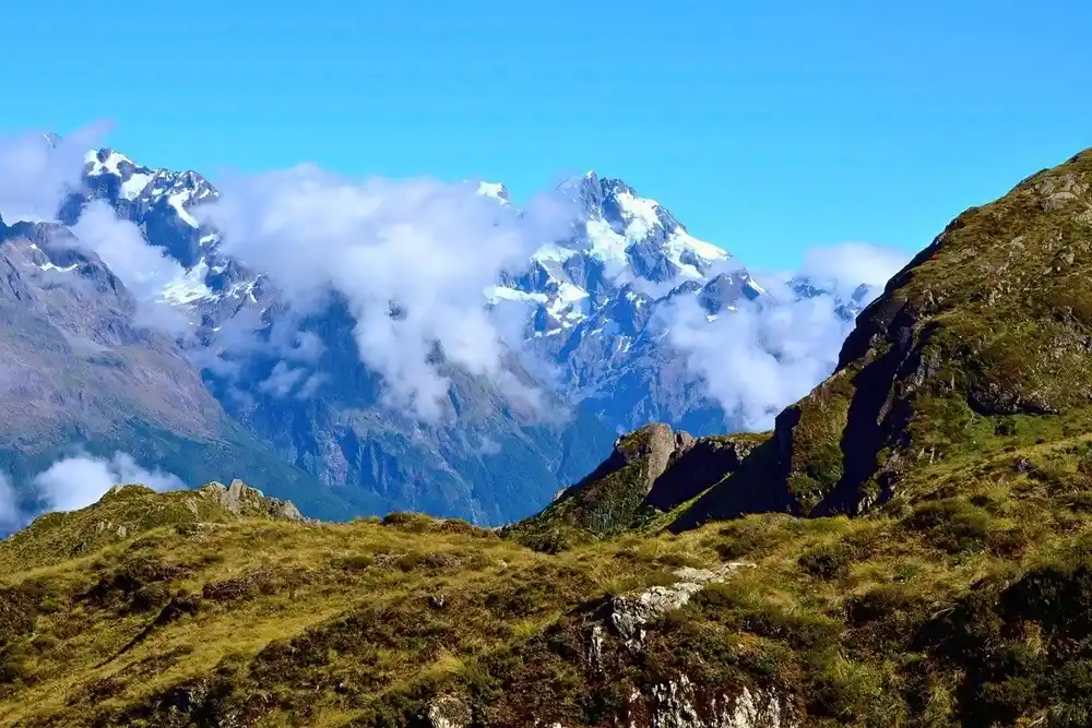 Snow-capped peaks of the Southern Alps in New Zealand rising above rocky hills and drifting clouds under a bright blue sky.