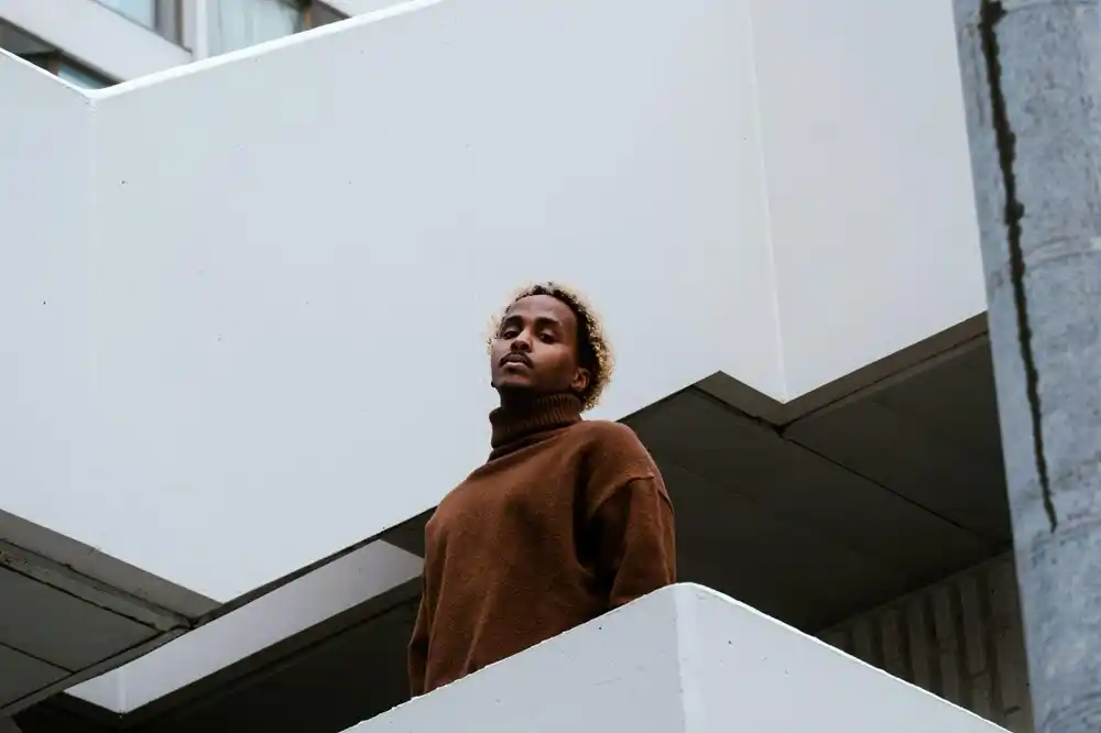 Man wearing a brown roll-neck jumper, standing on a modern building balcony.