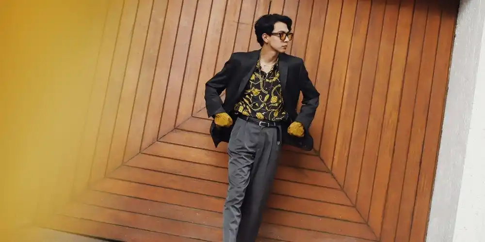 Man wearing a black blazer over a patterned shirt with grey trousers and loafers, leaning against a large wooden door.