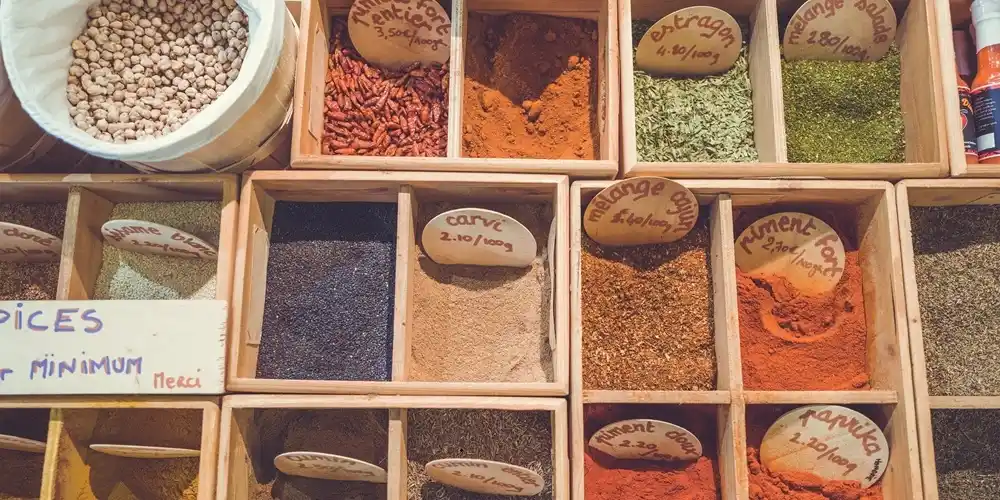 An assortment of colourful spices, herbs and dried ingredients displayed in wooden boxes at a market stall, with handwritten labels and prices.