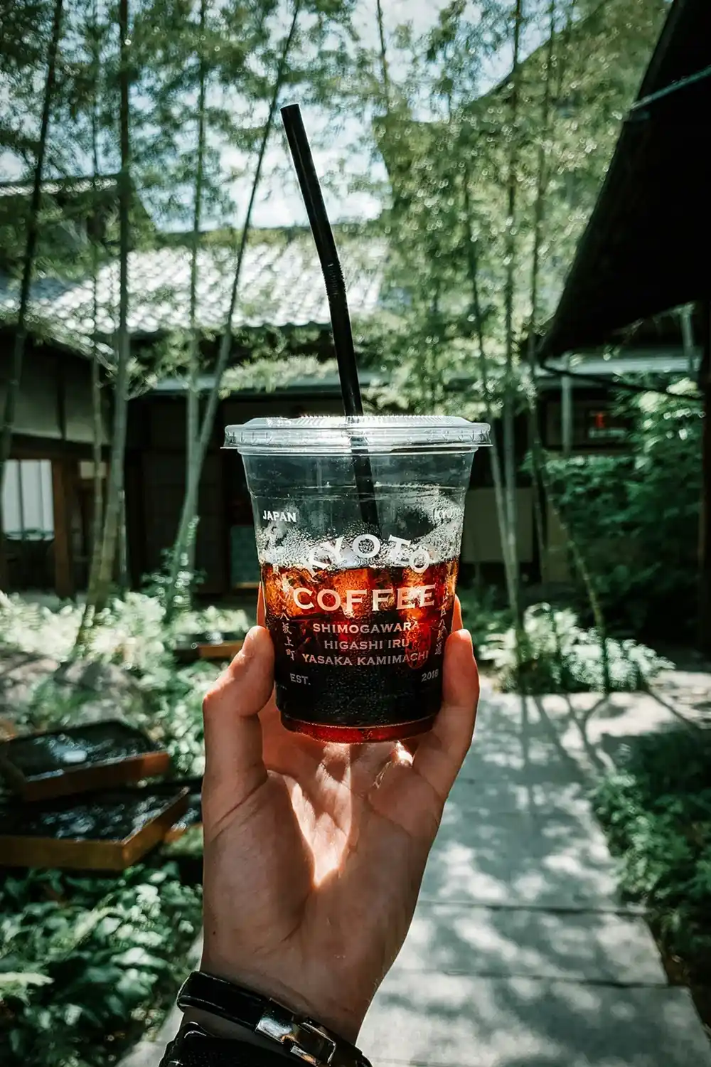 Hand holding an iced coffee beneath the trees at a Kyoto coffee shop.