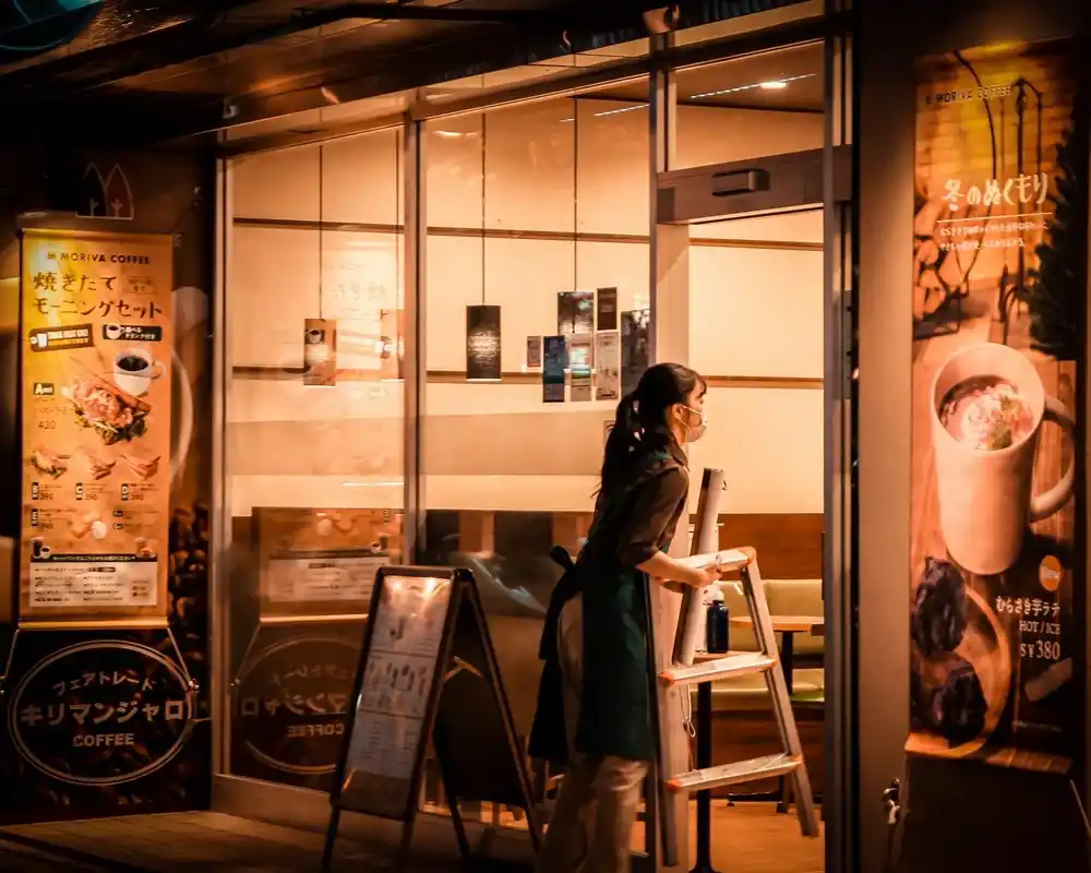 Staff member arranging equipment outside a coffee shop in Tokyo, surrounded by coffee signs and warm lighting.
