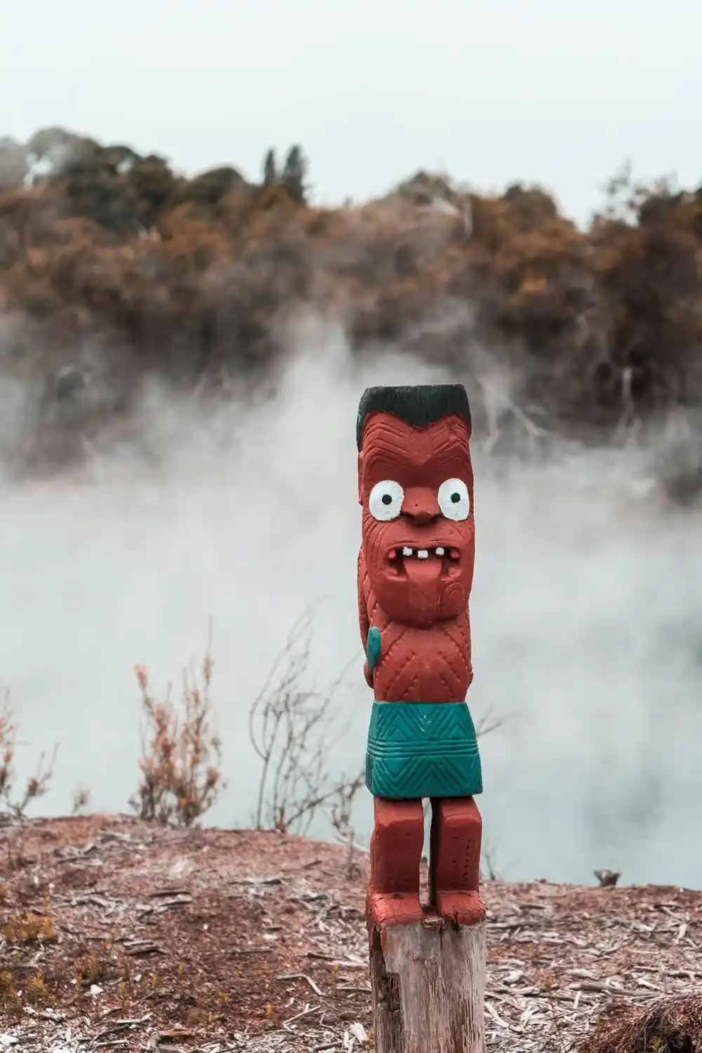 Red-painted carved wooden Māori figure with wide white eyes and protruding tongue, before a steaming geothermal lake.