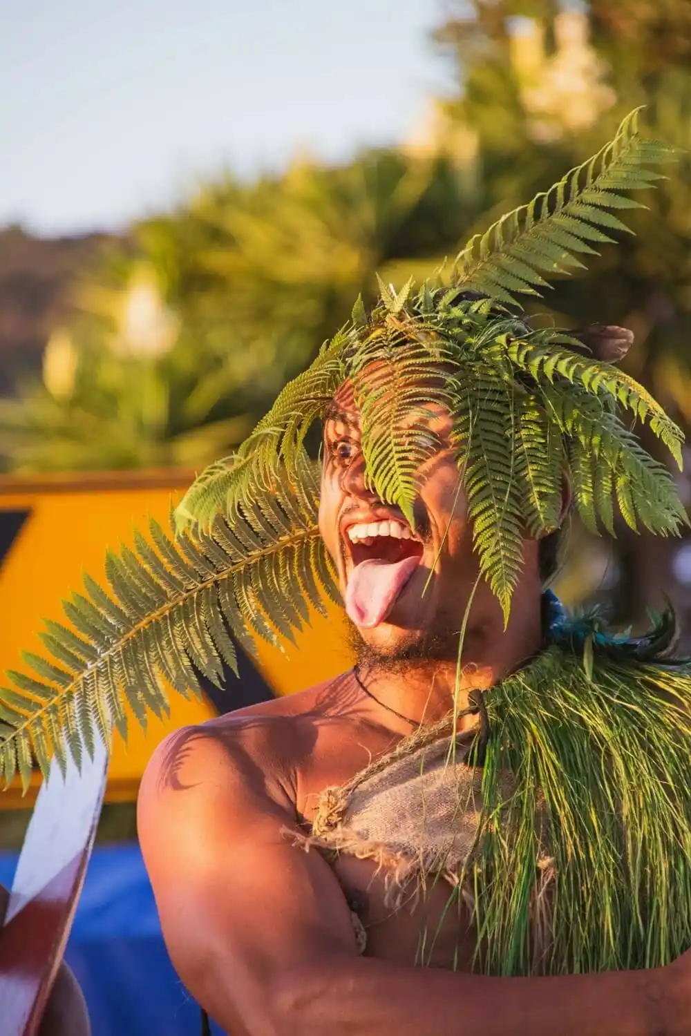 Māori haka performer in fern headdress and green fibre cape, eyes wide and tongue thrust out, lit by warm evening sun.