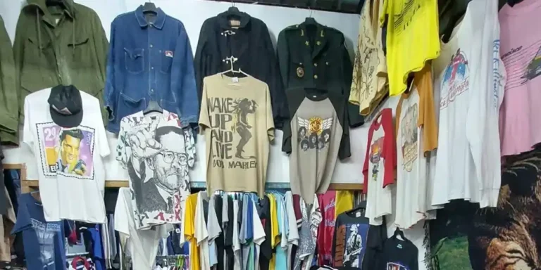 Crowded thrift stall with vintage band and graphic T-shirts, denim and military jackets on the walls and piles of folded tees.