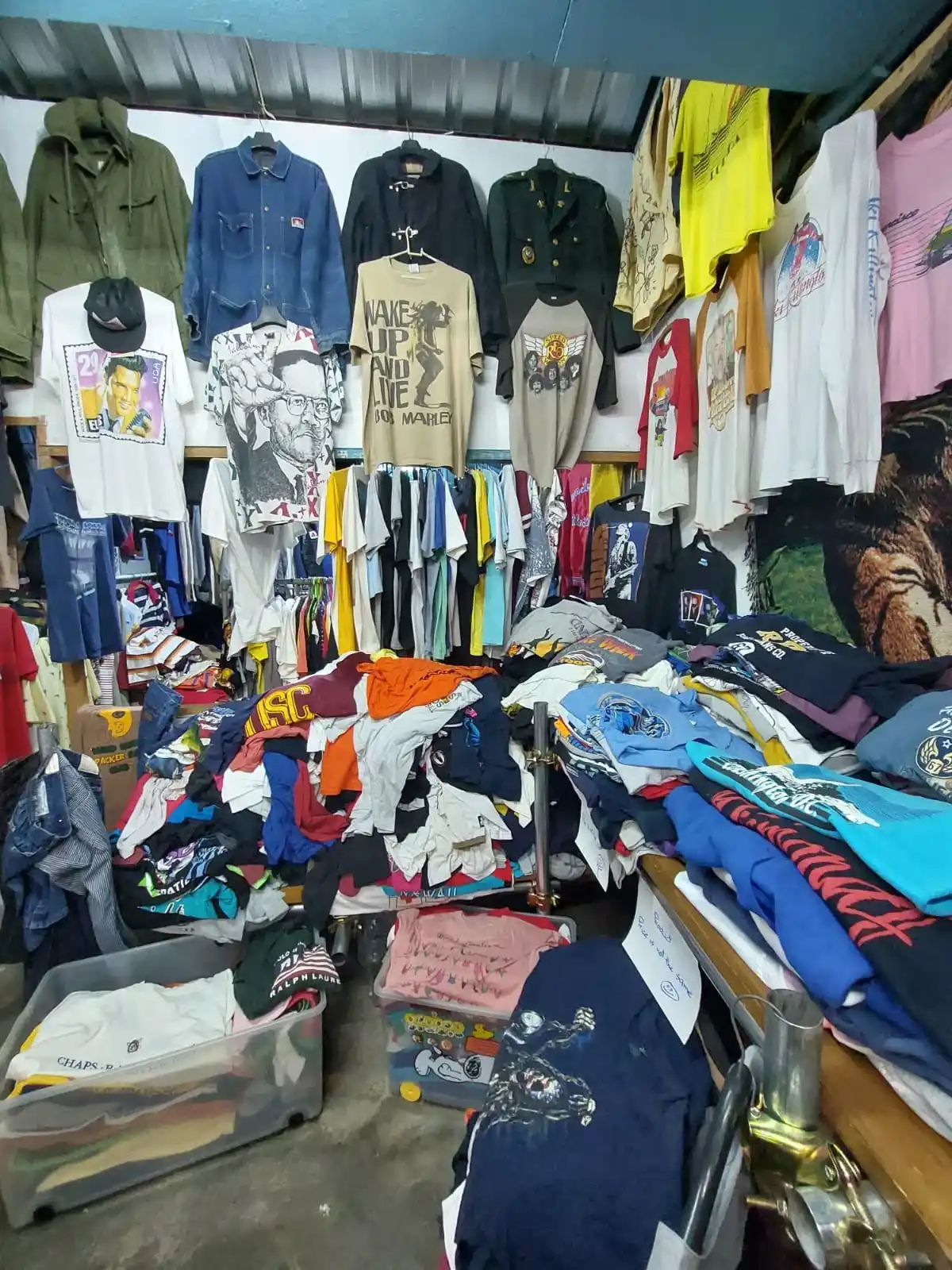 Crowded thrift stall with vintage band and graphic T-shirts, denim and military jackets on the walls and piles of folded tees.