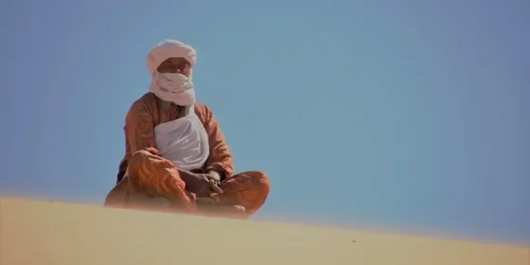 Tuareg man in a white turban and veil and rust-orange patterned robe sitting cross-legged on a sand dune under a blue sky.