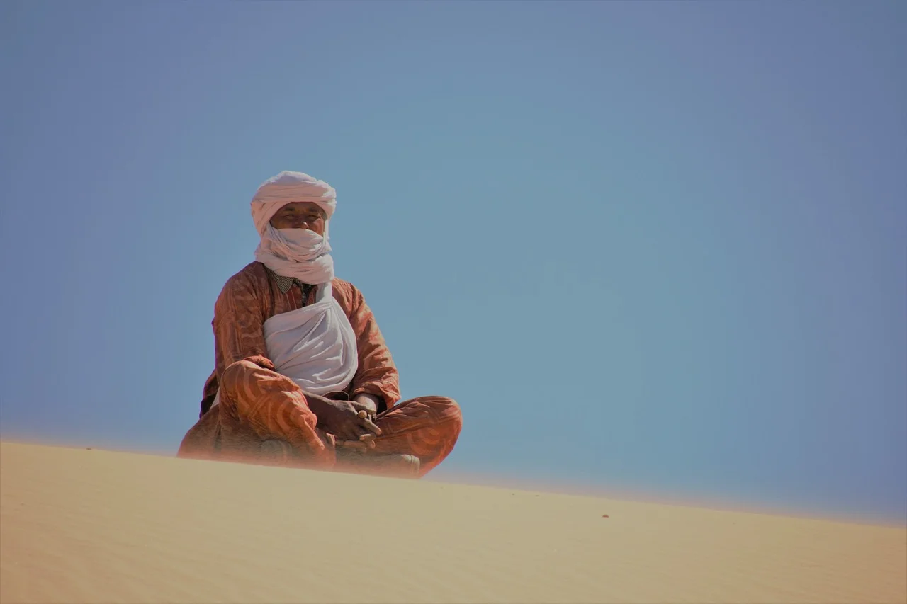 Tuareg man in a white turban and veil and rust-orange patterned robe sitting cross-legged on a sand dune under a blue sky.