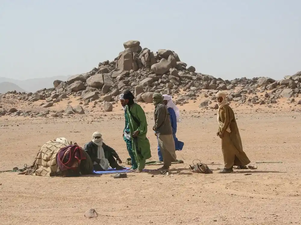A group of Tuareg men in turbans and long robes at a desert camp, one seated by saddlebags, a rocky outcrop behind.