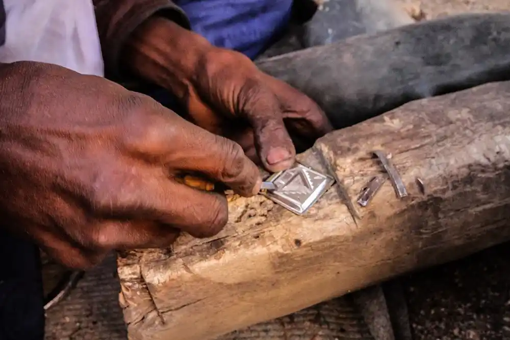 Close-up of a silversmith’s hands engraving a geometric pattern into a diamond-shaped silver pendant on a wooden block.