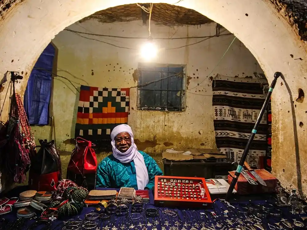 Trader in a white turban and teal robe behind a table of silver bracelets, pendants and rings, lit by a bare bulb.
