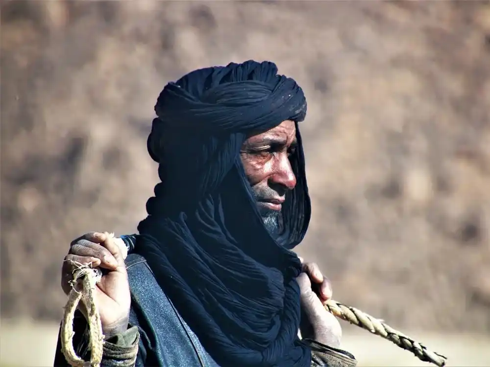 Tuareg man in a dark indigo turban and veil with a grey beard, holding a braided rope across his shoulders, in profile.