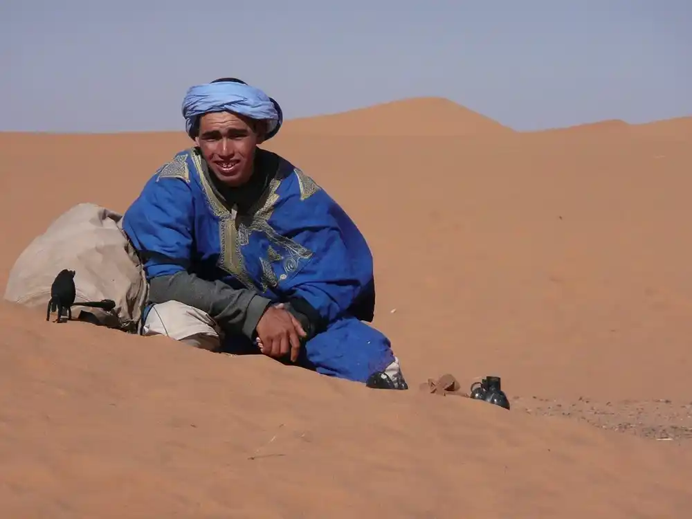 Smiling young man in a light-blue turban and gold-embroidered blue robe sitting on an orange sand dune beside a bag.