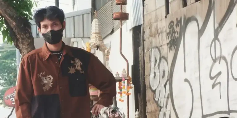 Man in a face mask, brown patchwork overshirt with embroidery and plaid trousers, leaning on a metal barrier by graffiti.