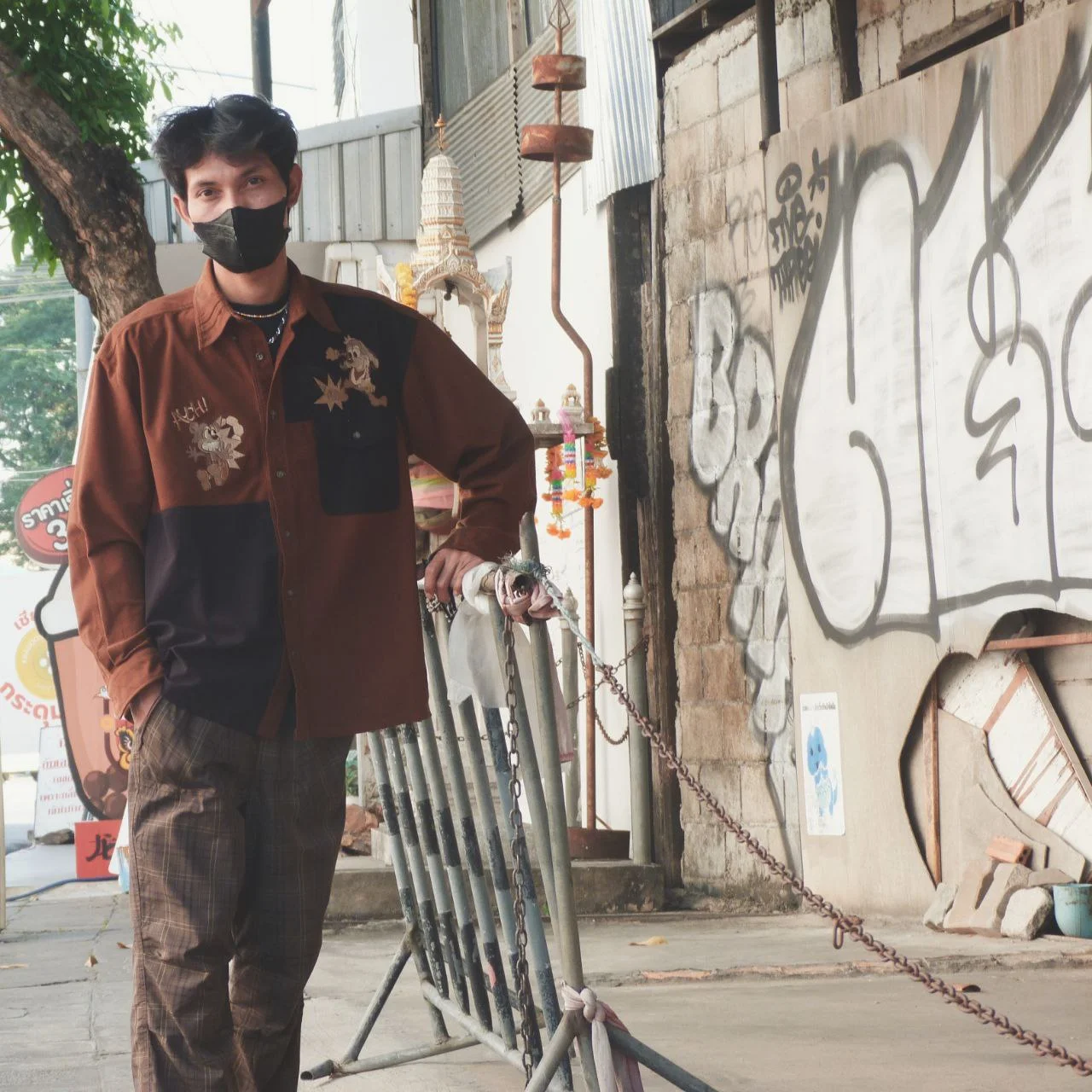 Man in a face mask, brown patchwork overshirt with embroidery and plaid trousers, leaning on a metal barrier by graffiti.